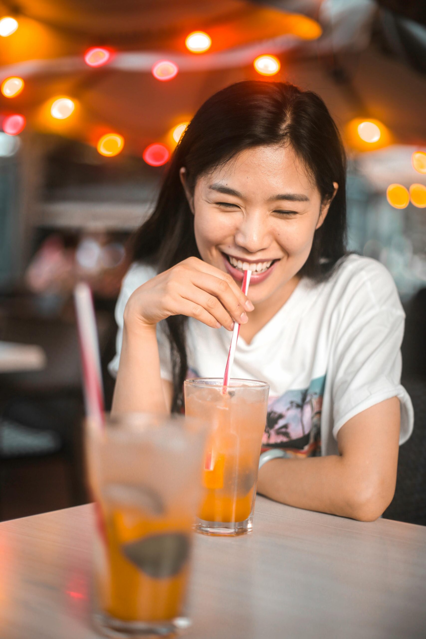 Happy young woman sipping a refreshing drink at a lively café with colorful lights and a cheerful atmosphere.